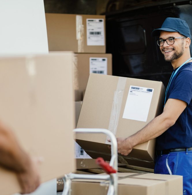 Young happy manual worker carrying cardboard boxes in delivery van while communicating with his colleagues.
