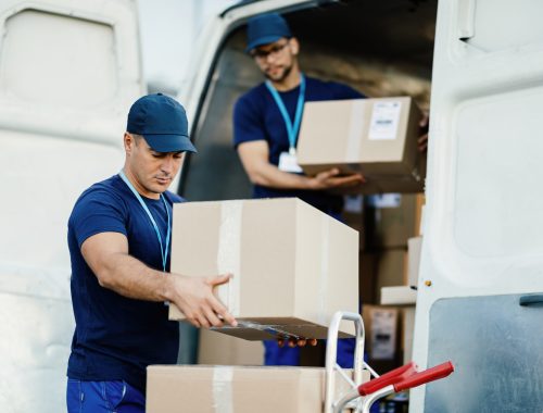 Young courier and his colleague unloading cardboard boxes from delivery van.